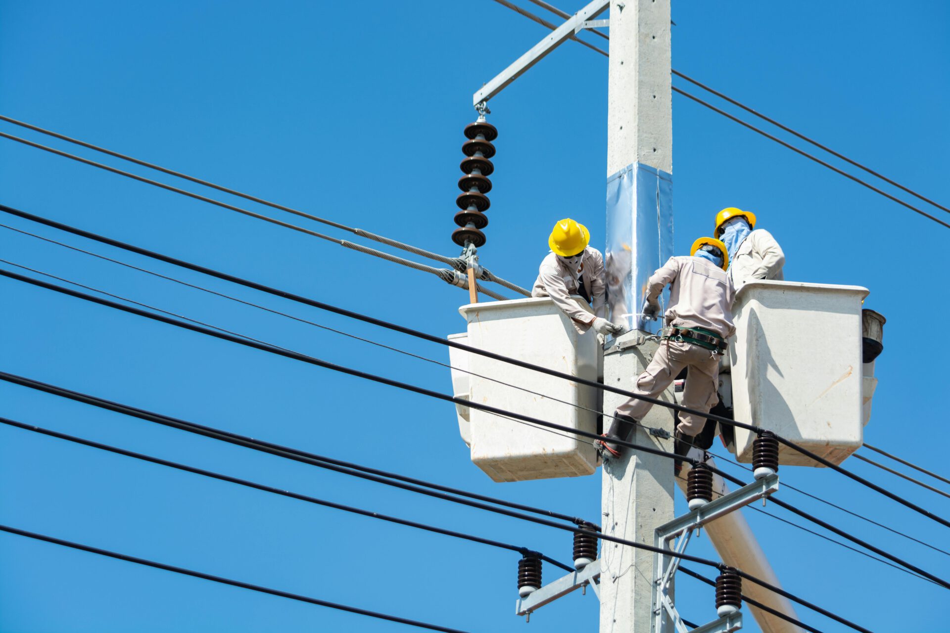 Electricians wearing safety gear work on power lines using lifts against a clear blue sky.